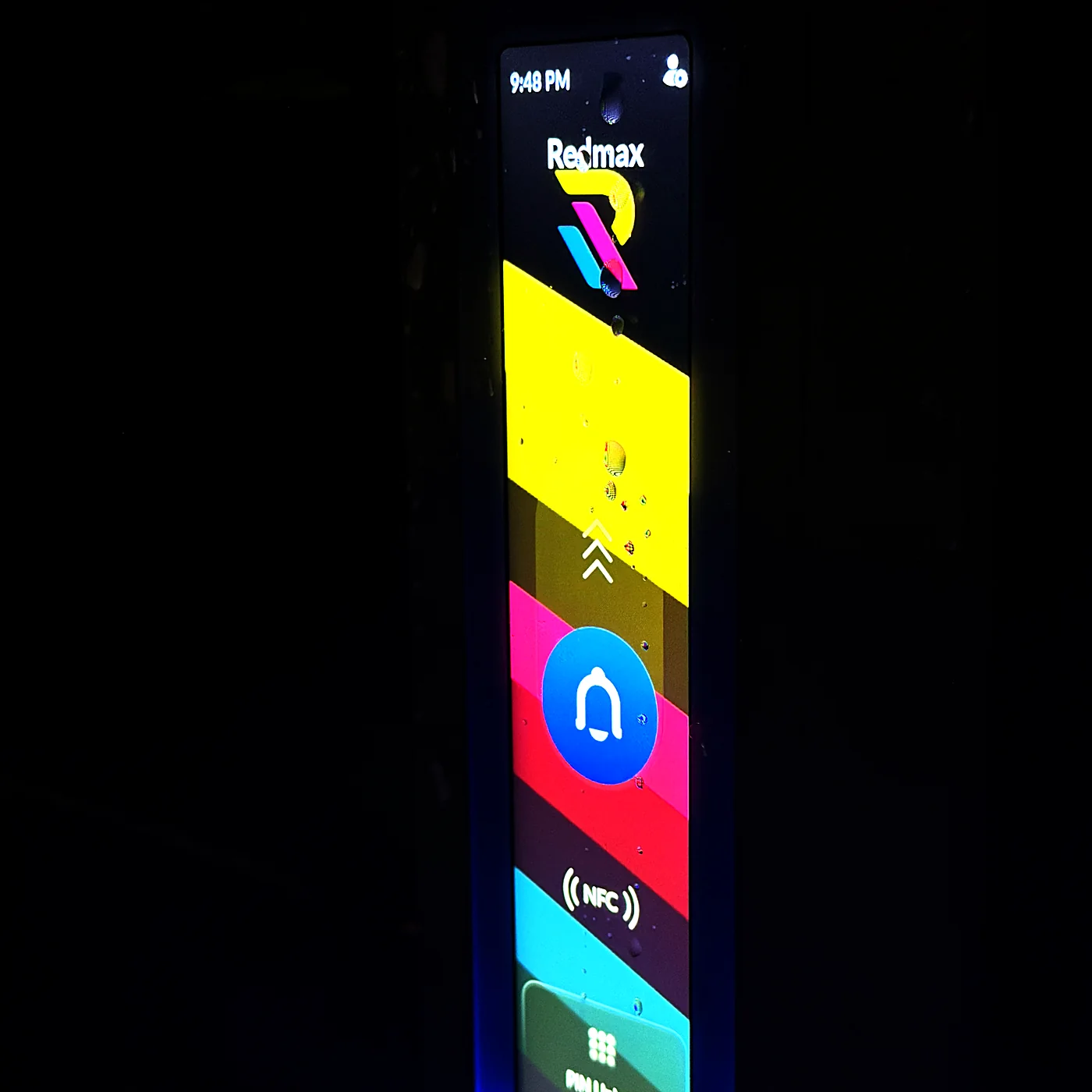 A tall touchscreen door intercom lit up against a dark exterior wall at night, showing a tenant directory and a call button.