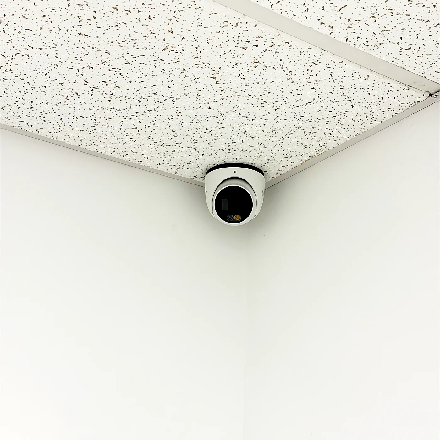 A white dome camera mounted in the corner where a suspended ceiling meets two white walls.