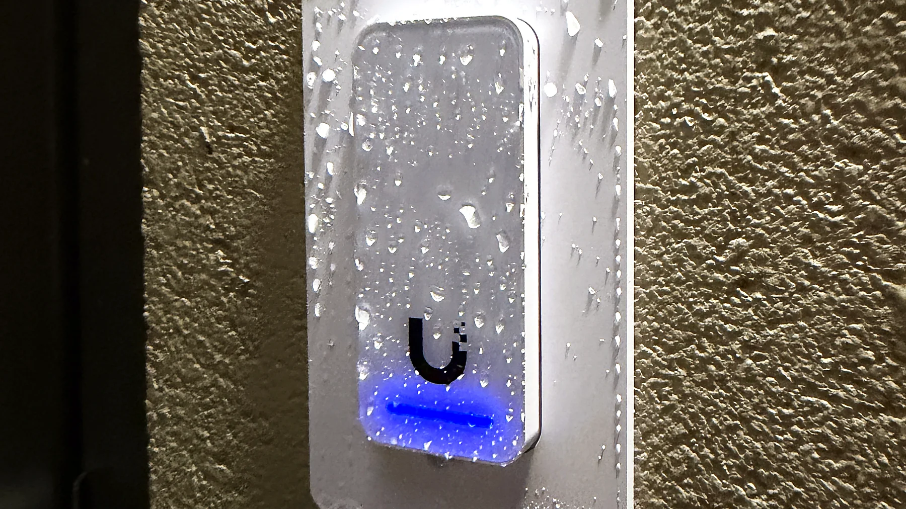 An access control reader mounted on an exterior block wall at night, beaded with rain, its indicator lit blue.