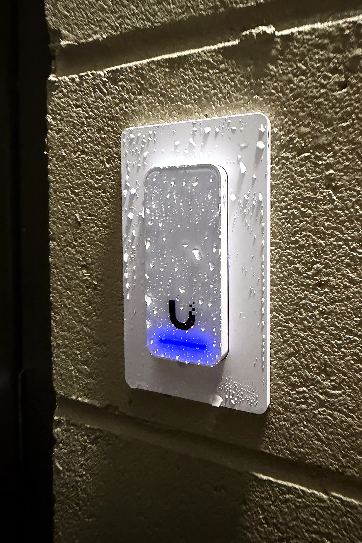 An access control reader mounted on an exterior block wall at night, beaded with rain, its indicator lit blue.