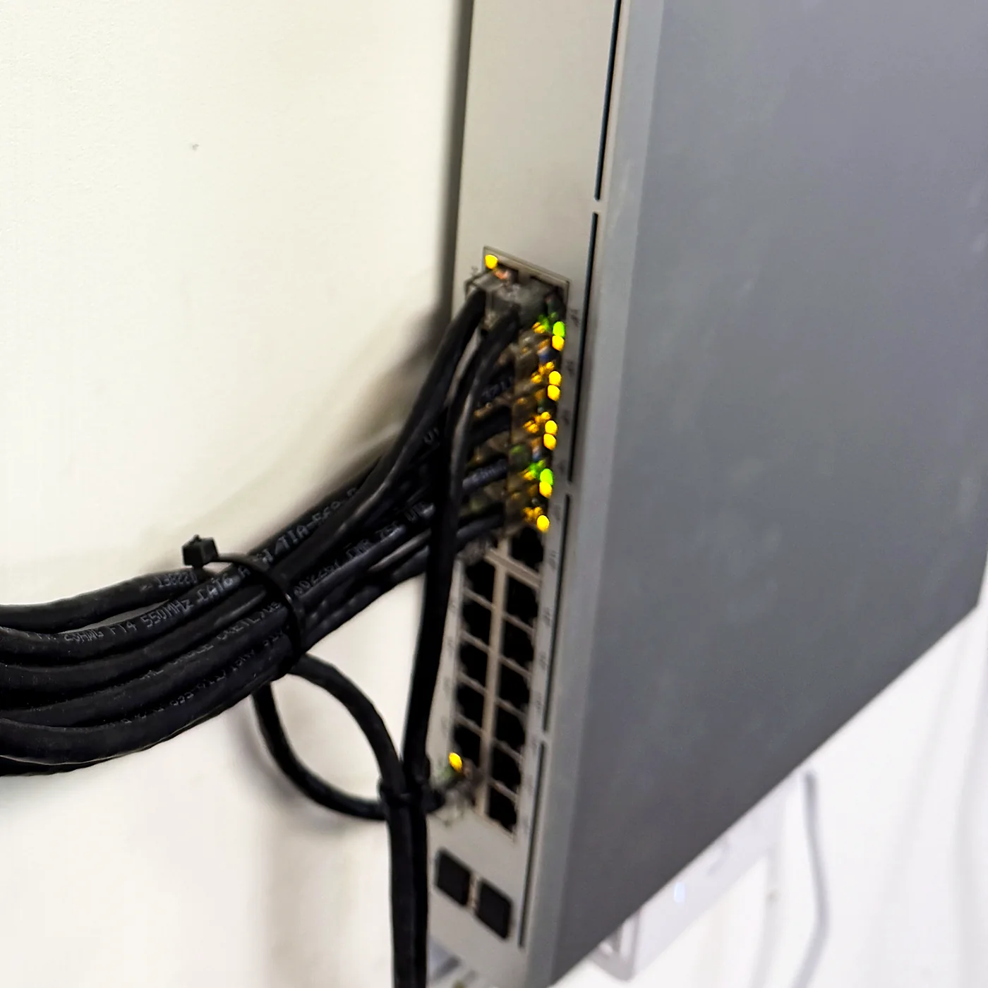 A wall-mounted network switch with a bundle of black patch cables sweeping in from the left in a single dressed arc into the switch ports, lit link LEDs along the top row, against a clean white wall.
