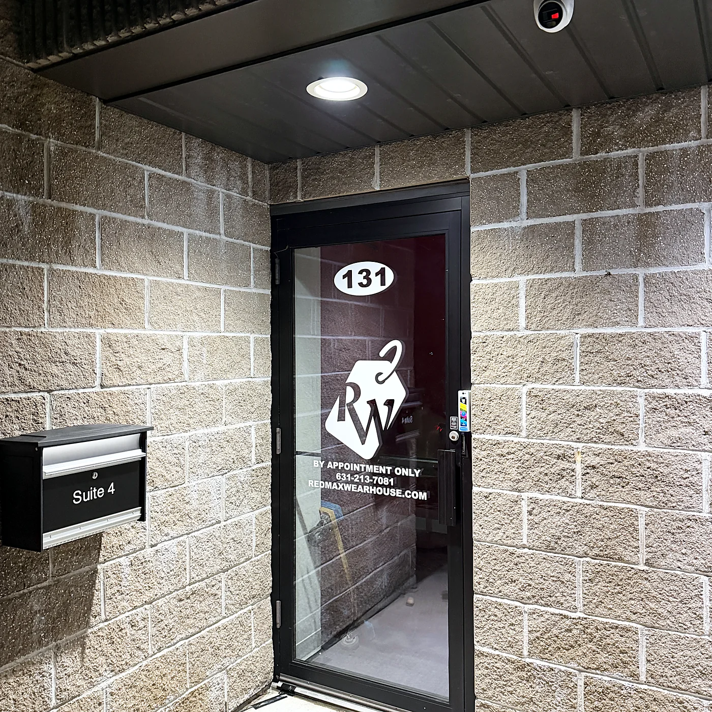A camera mounted above a glass entry door of a commercial suite at night, the block wall lit by a recessed downlight.