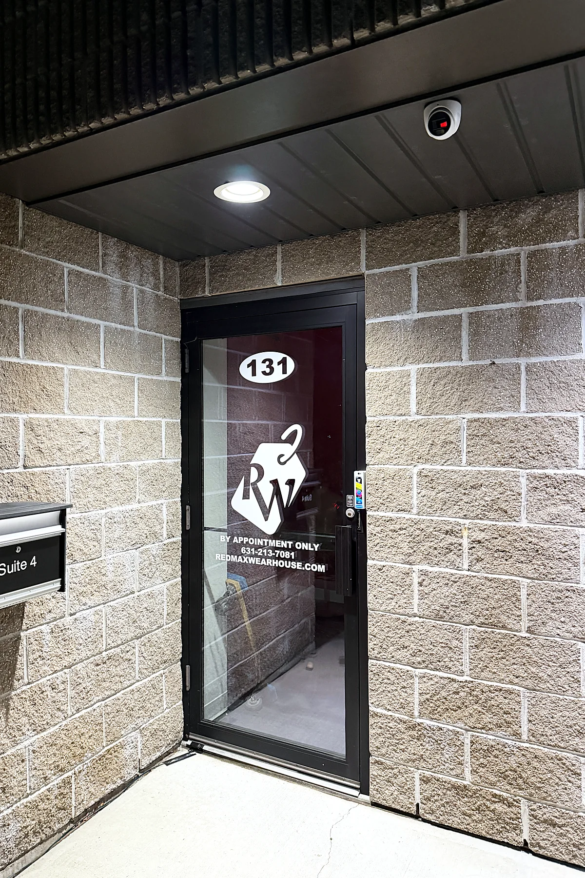 A camera mounted above a glass entry door of a commercial suite at night, the block wall lit by a recessed downlight.