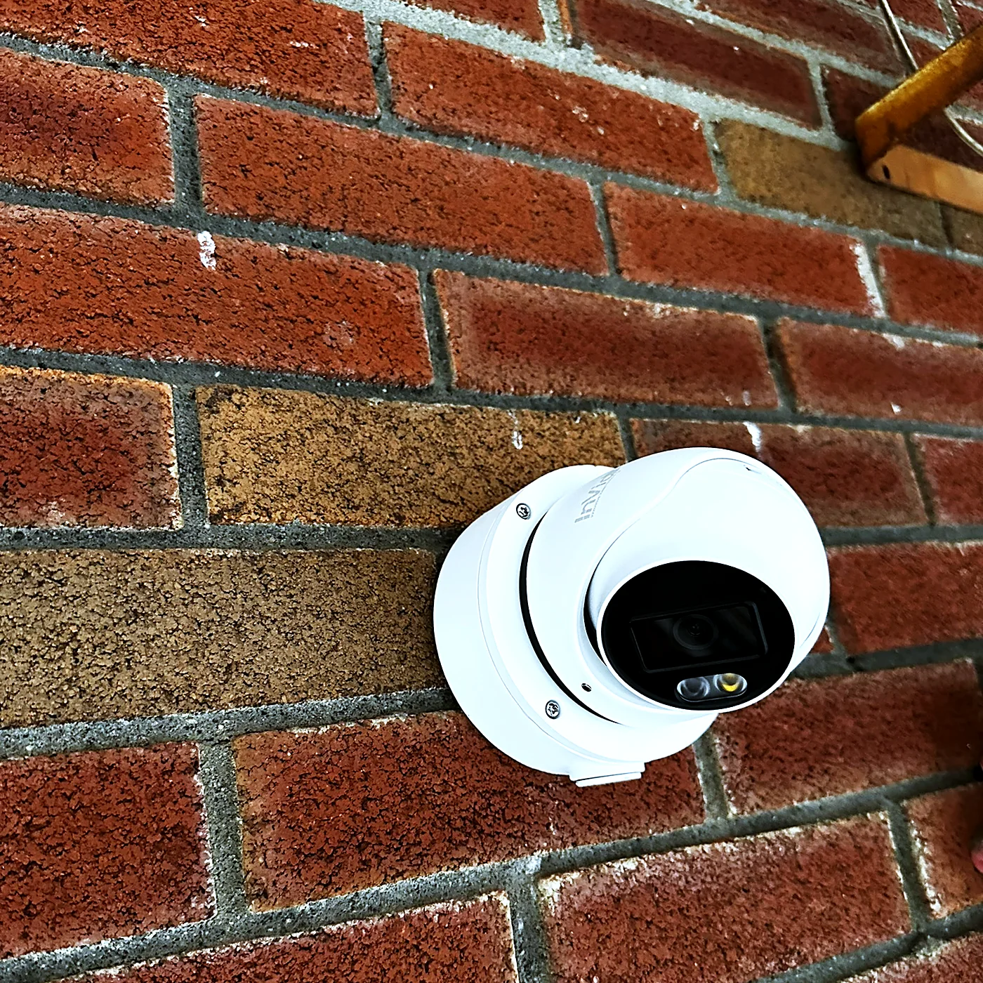 A white turret camera mounted on a red brick wall in direct sunlight, aimed down and to the left.