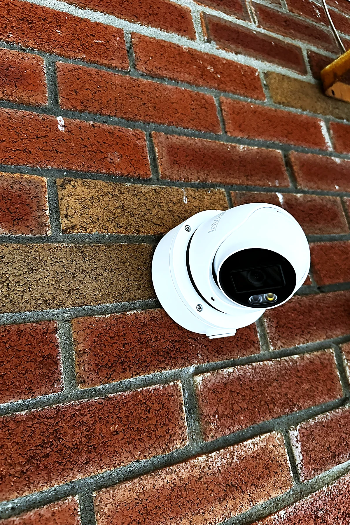 A white turret camera mounted on a red brick wall in direct sunlight, aimed down and to the left.
