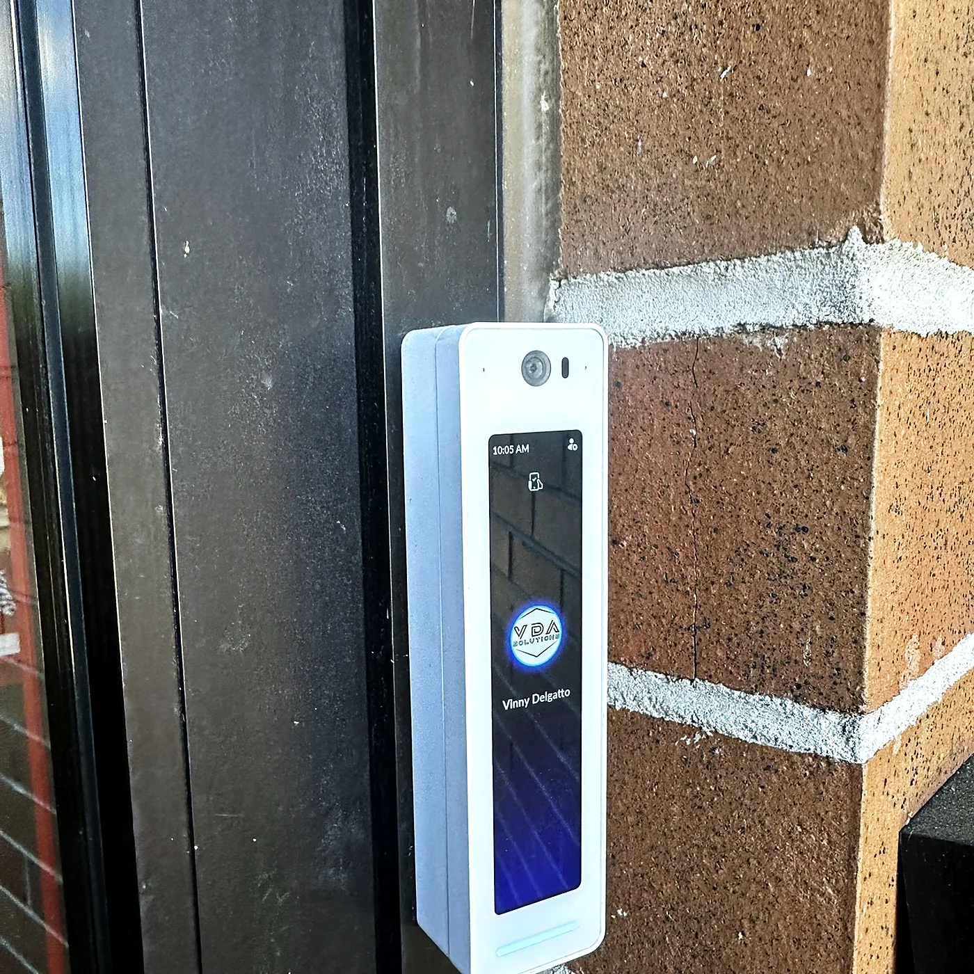 A tall touchscreen door intercom mounted on a brick pier beside a dark storefront frame, showing a name and a call button.