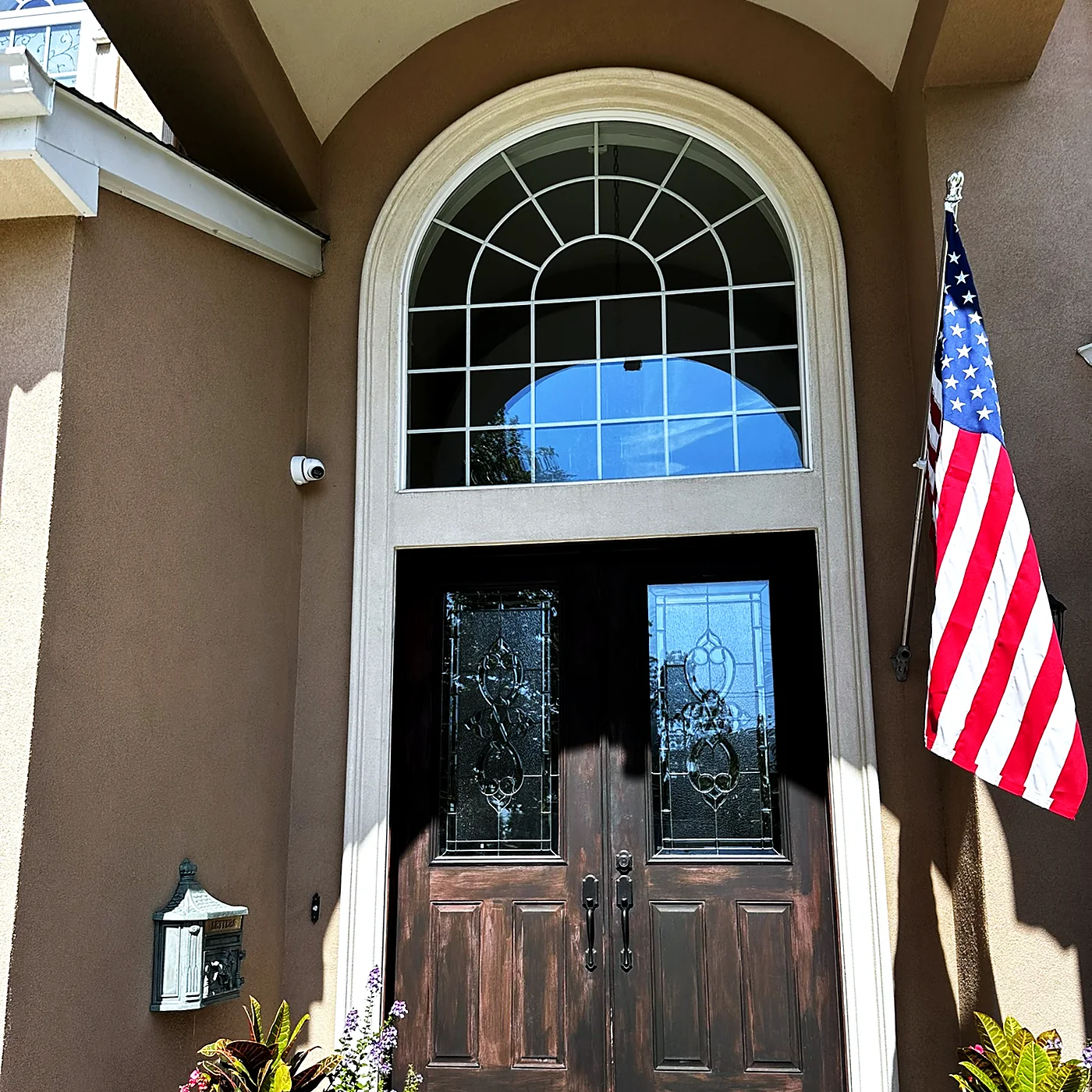 A camera mounted high on the stucco elevation of a house beside an arched entry window and a flag.