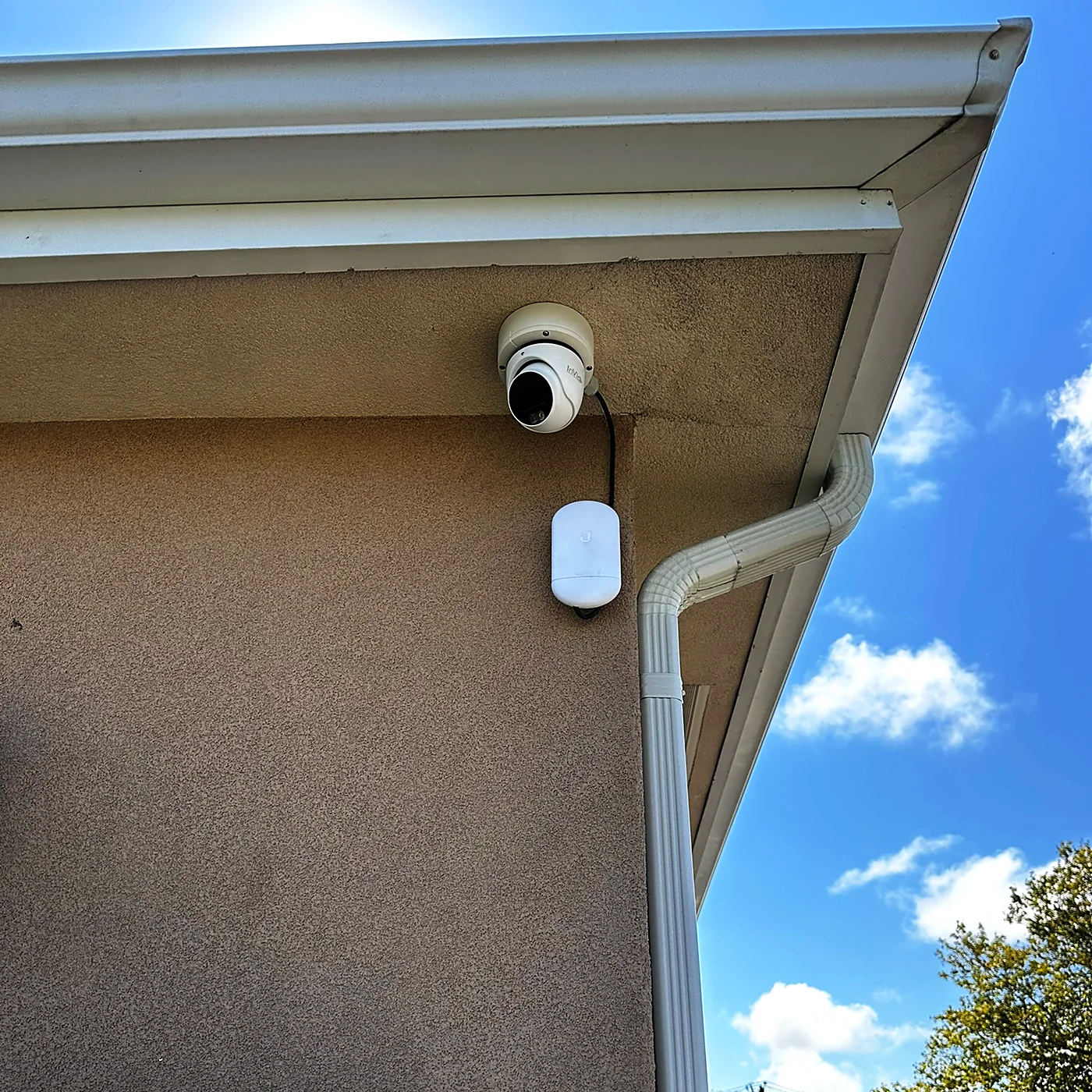 A camera and a small weatherproof junction box mounted under a stucco soffit, blue sky and clouds behind.