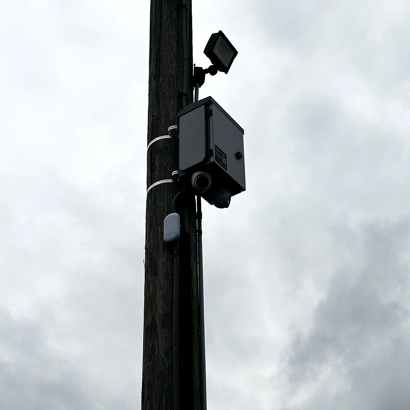 A camera and a weatherproof enclosure mounted on a timber pole in a boat yard, overcast sky behind.