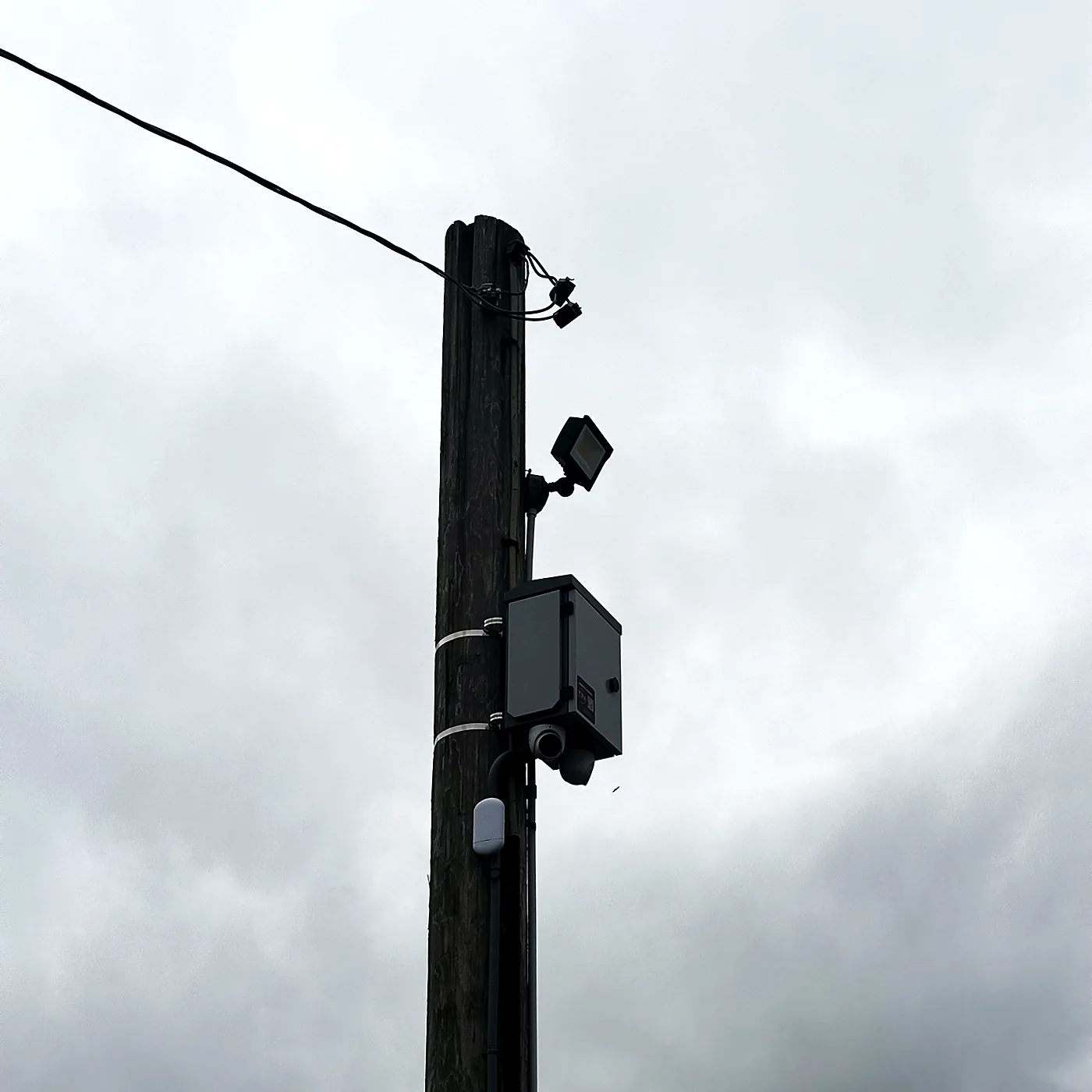 A camera and a weatherproof enclosure mounted partway up a utility pole against an overcast sky.