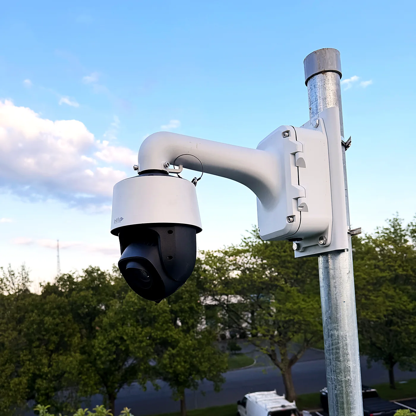 A pan-tilt-zoom camera on a galvanised pole mount against a blue sky, a parking lot visible below.