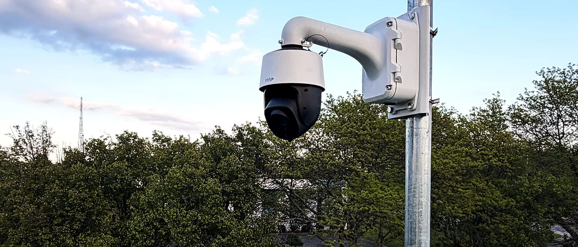 A pan-tilt-zoom camera and a weather-rated speaker mounted on the same pole above a treeline at dusk.