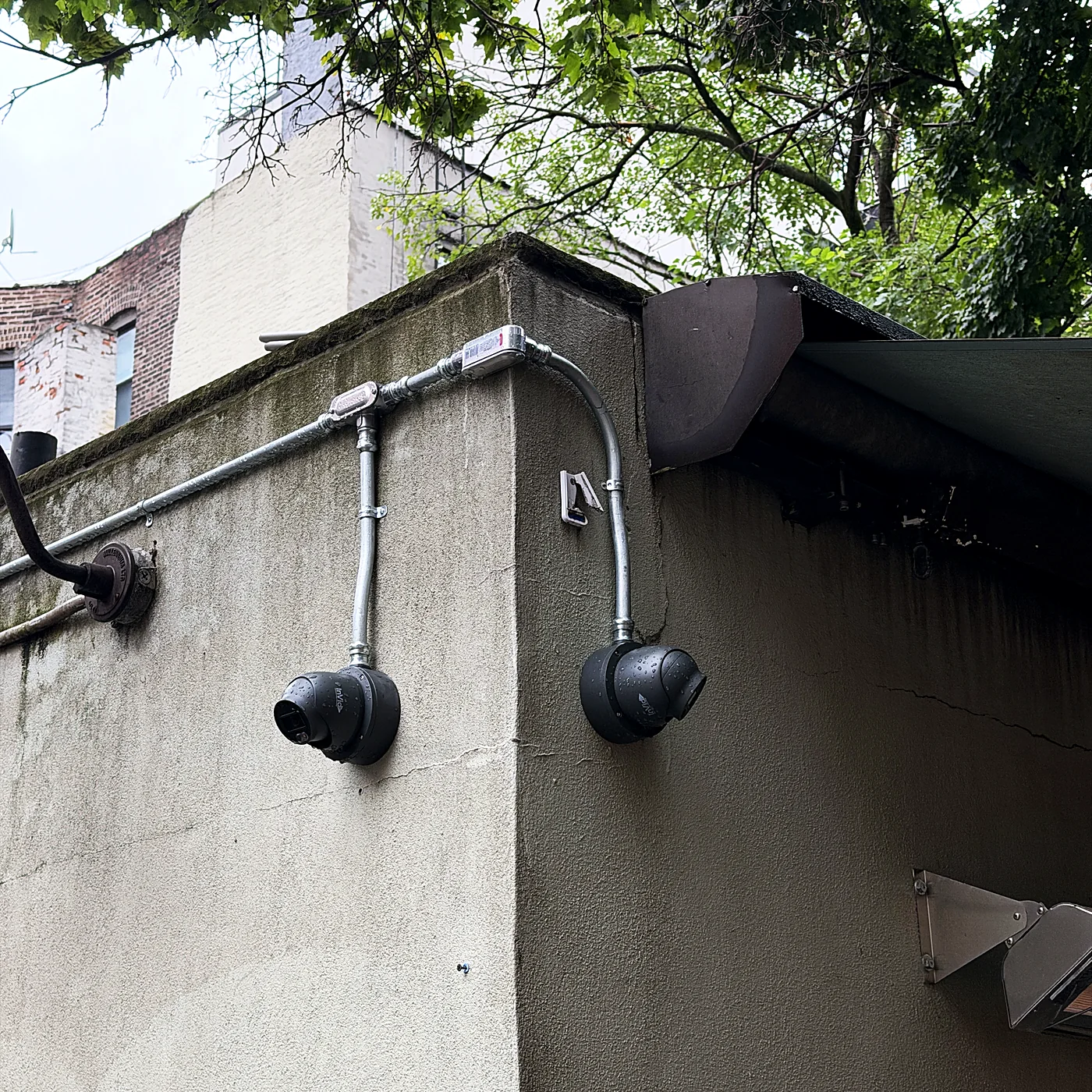 Two weather-rated pendant speakers mounted on a stucco wall, fed by exposed rigid conduit running along the parapet.