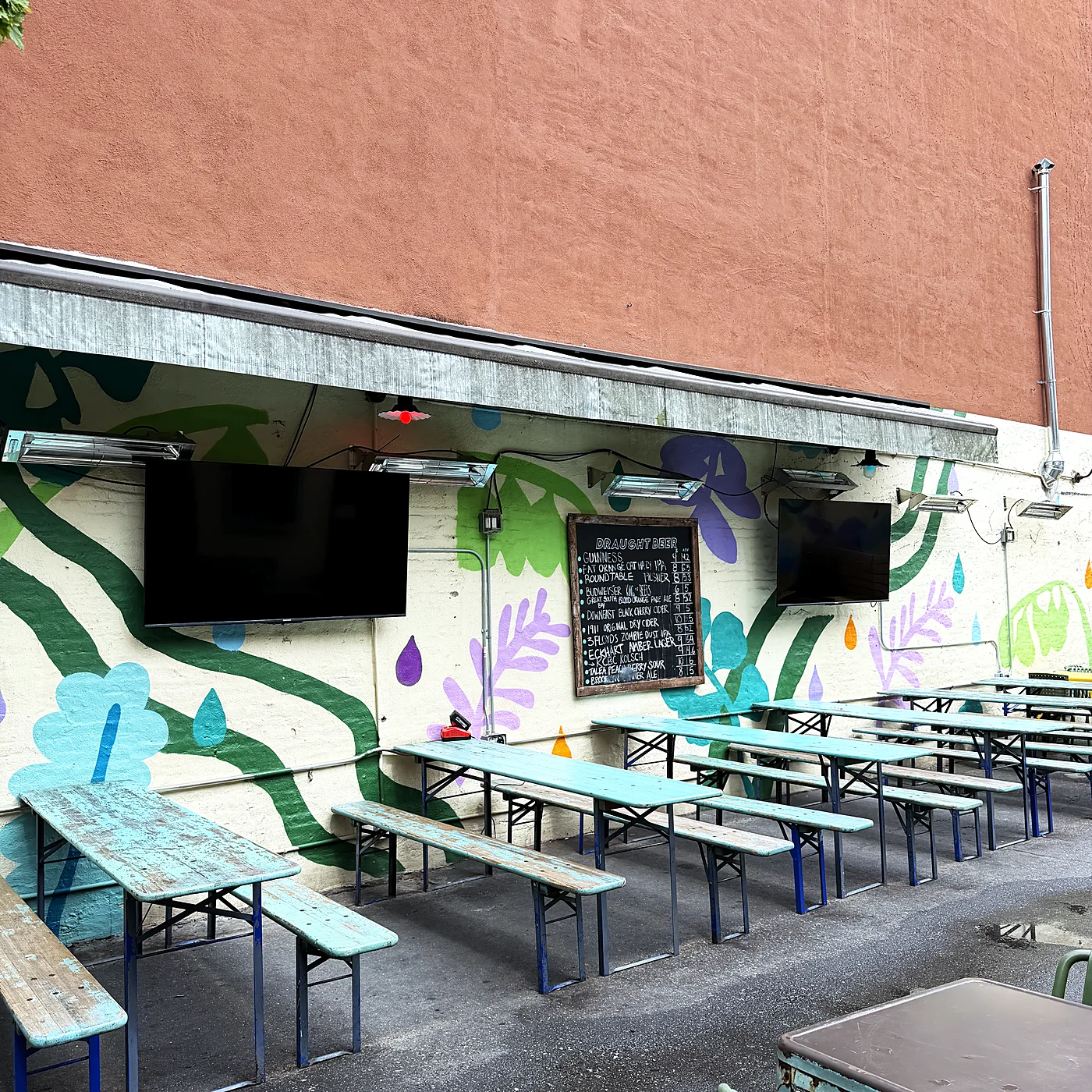 An outdoor patio with a painted mural, two televisions mounted under the roof overhang, and picnic benches below.