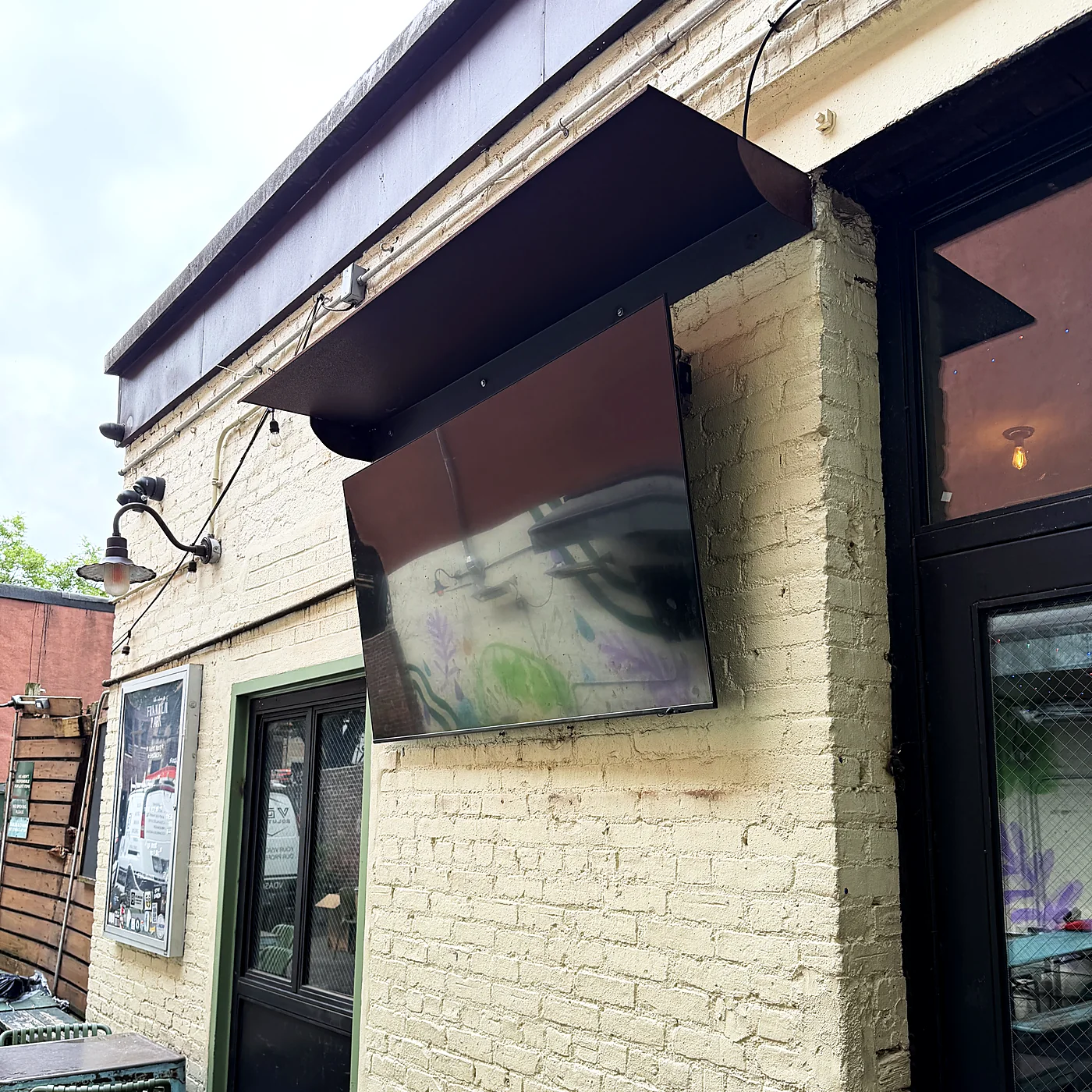 A television mounted under a metal awning on a painted brick exterior, with patio seating below.