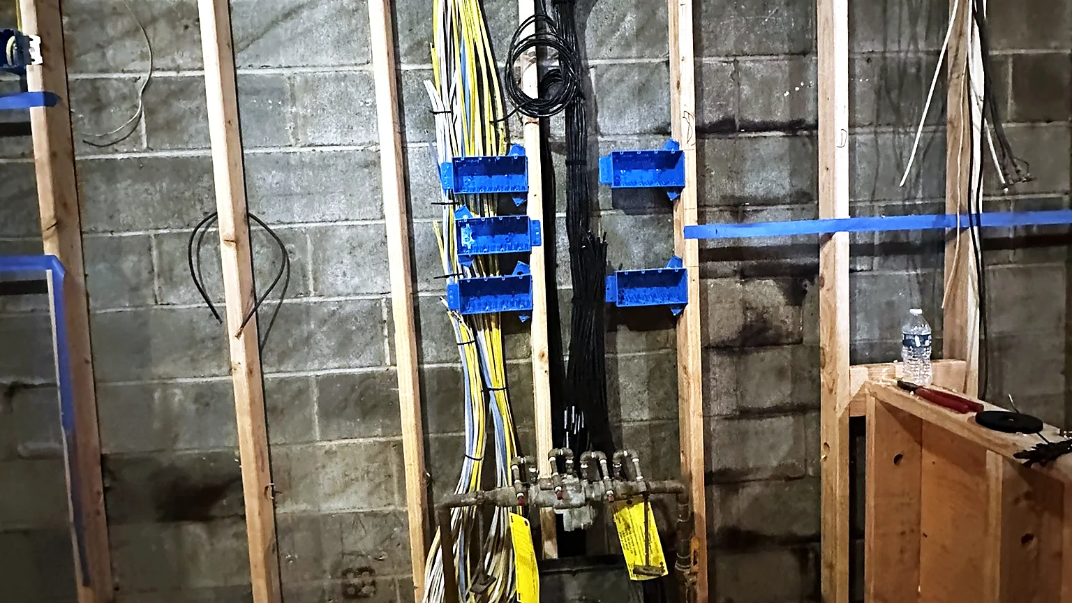 Cable bundles run vertically down a block wall between timber studs, supported on blue cable hangers, with a distribution point terminated at the base.