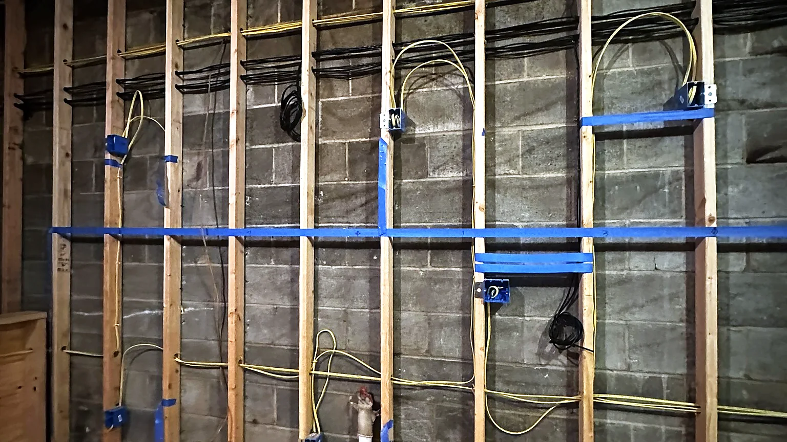A framed basement wall before drywall, with horizontal and vertical cable runs supported on blue hangers and device boxes set at each stud bay.