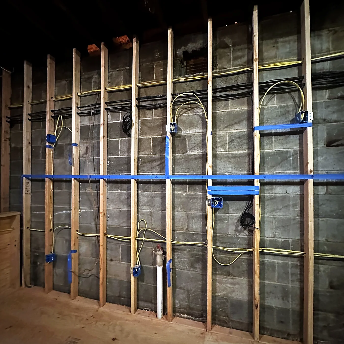 A framed basement wall before drywall, with horizontal and vertical cable runs supported on blue hangers and device boxes set at each stud bay.