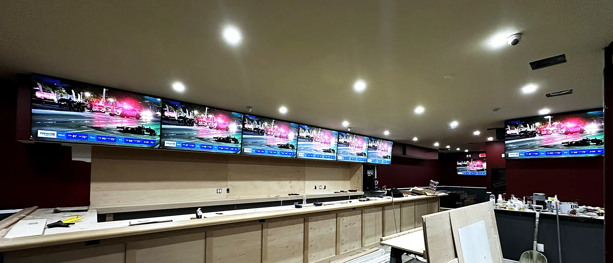 A long row of televisions mounted above a bar, all running live content, in a finished room with recessed lighting.