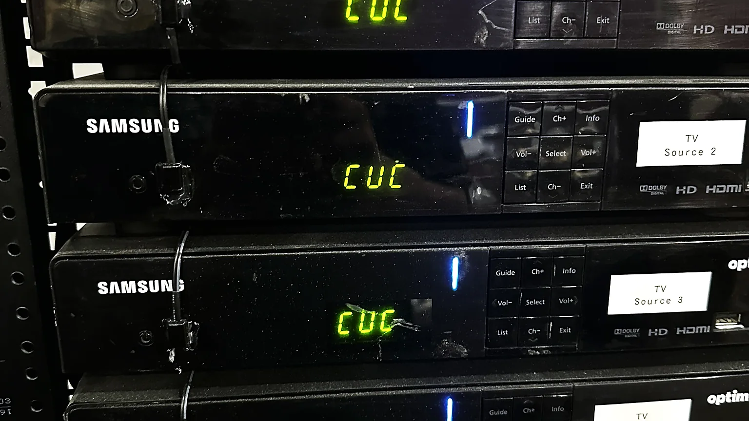 Four rack-mounted satellite tuners, each with a printed label reading TV Source and a number, stacked in an equipment rack.