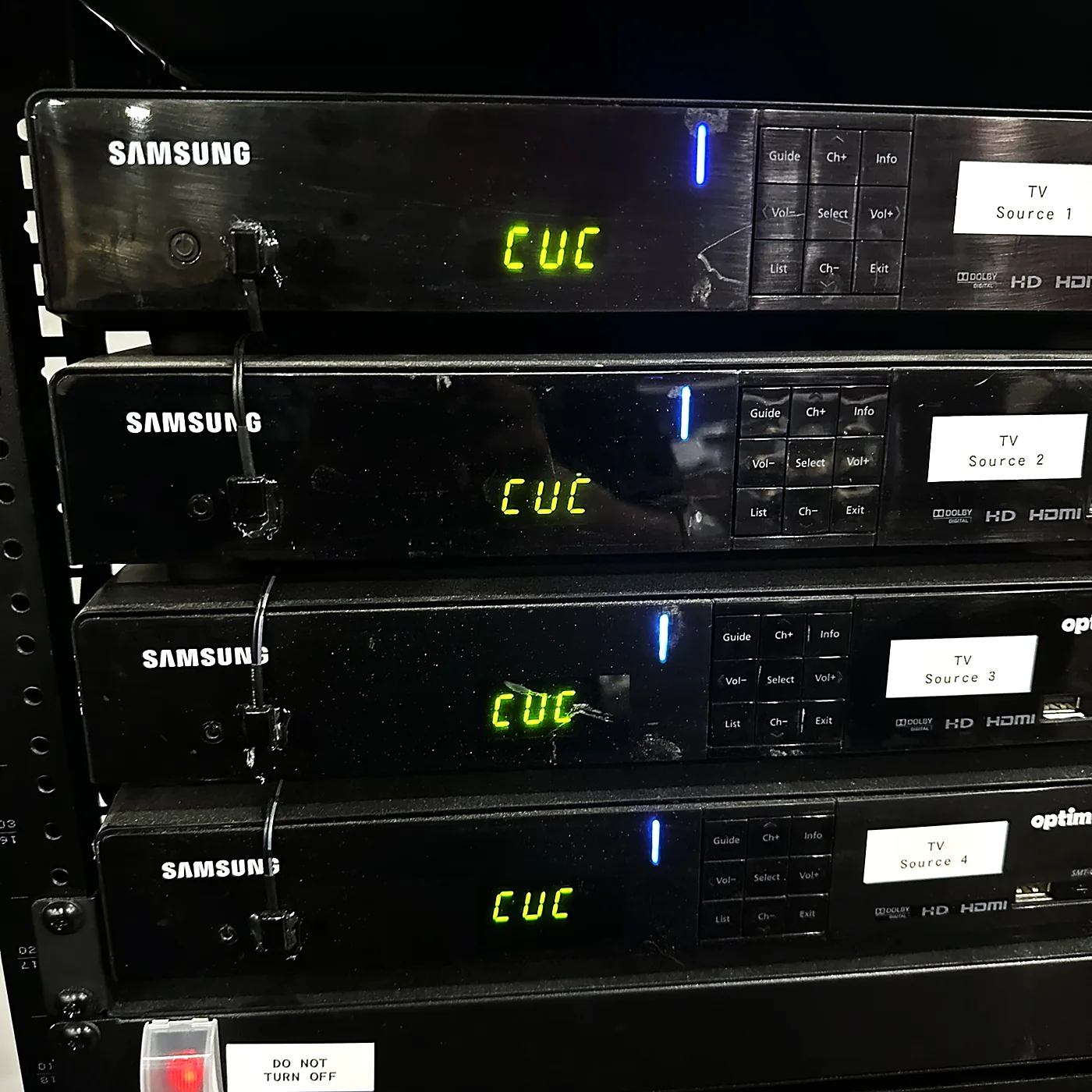Four rack-mounted satellite tuners, each with a printed label reading TV Source and a number, stacked in an equipment rack.