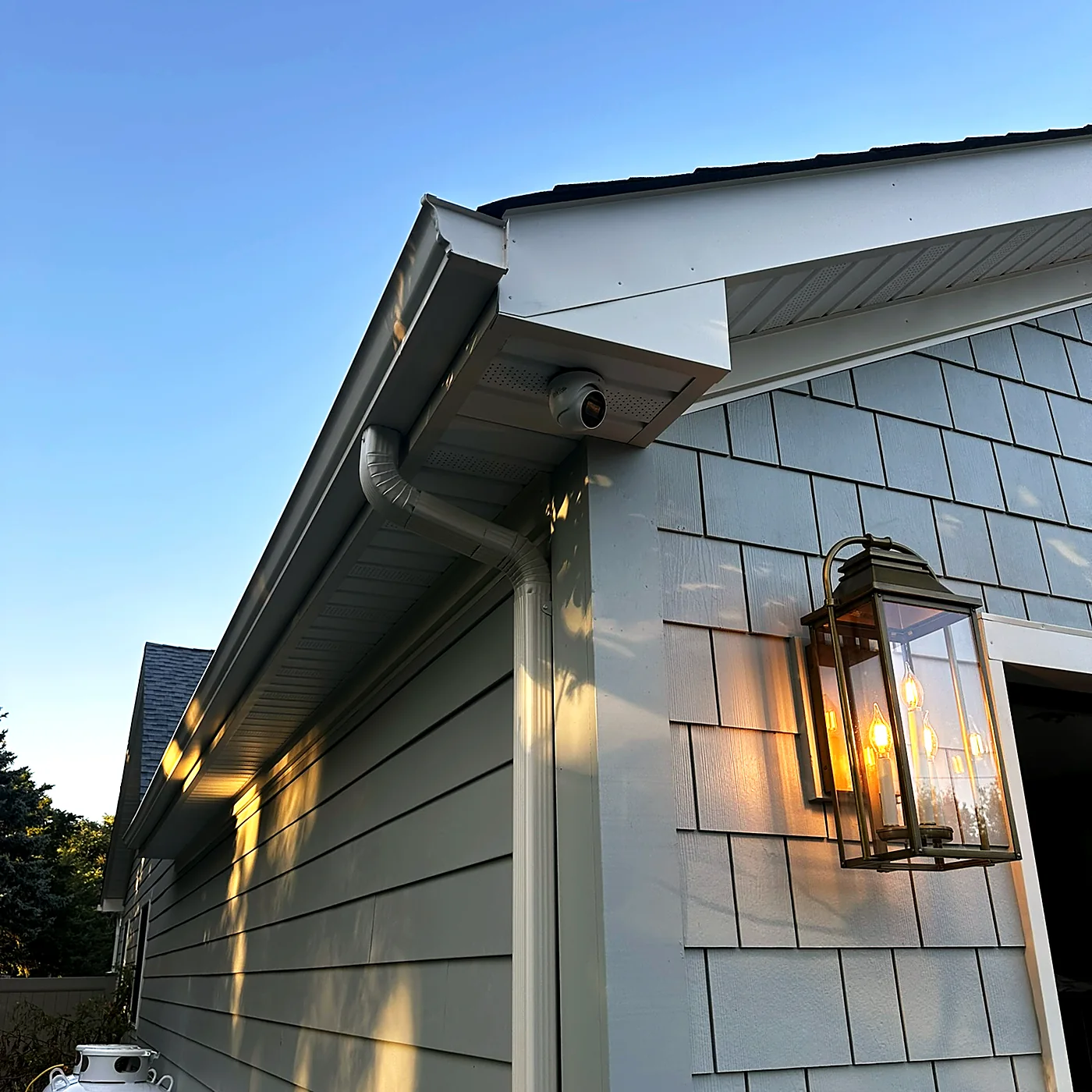A camera under the eave of a cedar-shingled house at dusk, a lantern sconce lit warm on the wall below.