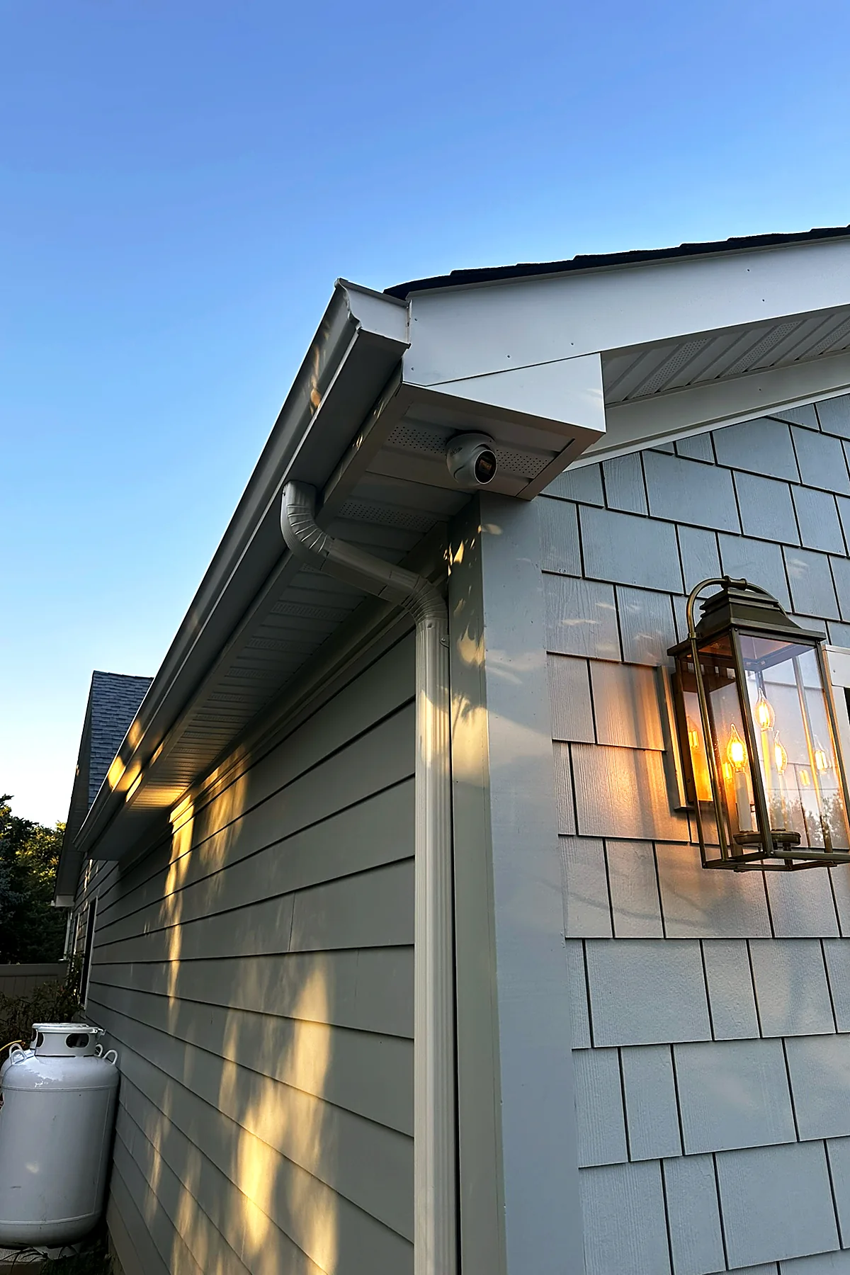 A camera under the eave of a cedar-shingled house at dusk, a lantern sconce lit warm on the wall below.