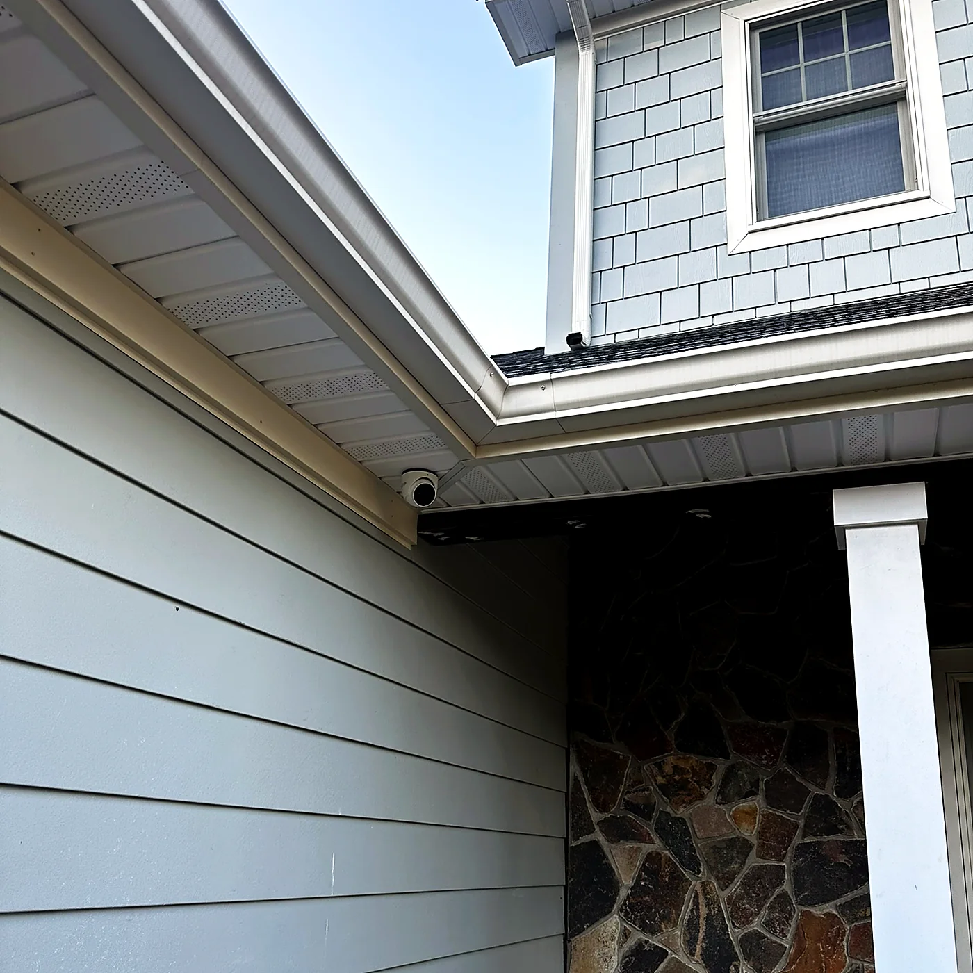 A camera mounted under the eave of a shingled house above a stone-clad entry column.
